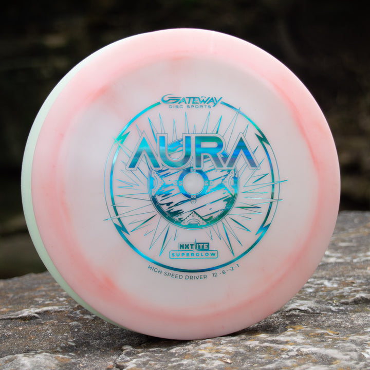 The Aura - NXT Lite Superglow, a white and light pink premium driver with blue artwork and "AURA" plus "GATEWAY DISC SPORTS" text, rests on a rock outdoors—ideal for smooth hyzer flips and ultimate glide.