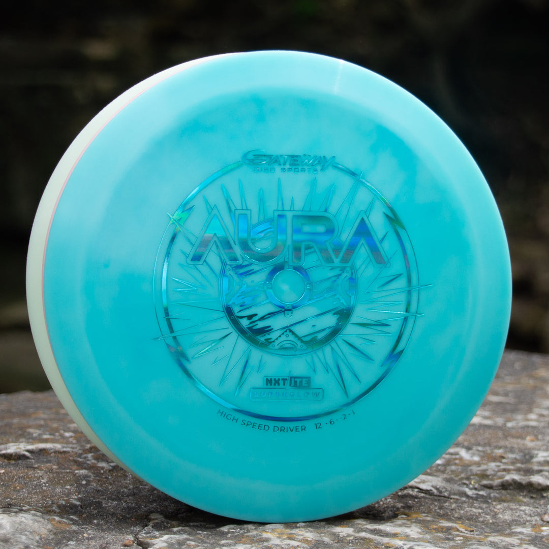 A vibrant NXT Lite Superglow Aura driver with starburst design rests on a rock—perfect for hyzer flips on the course.