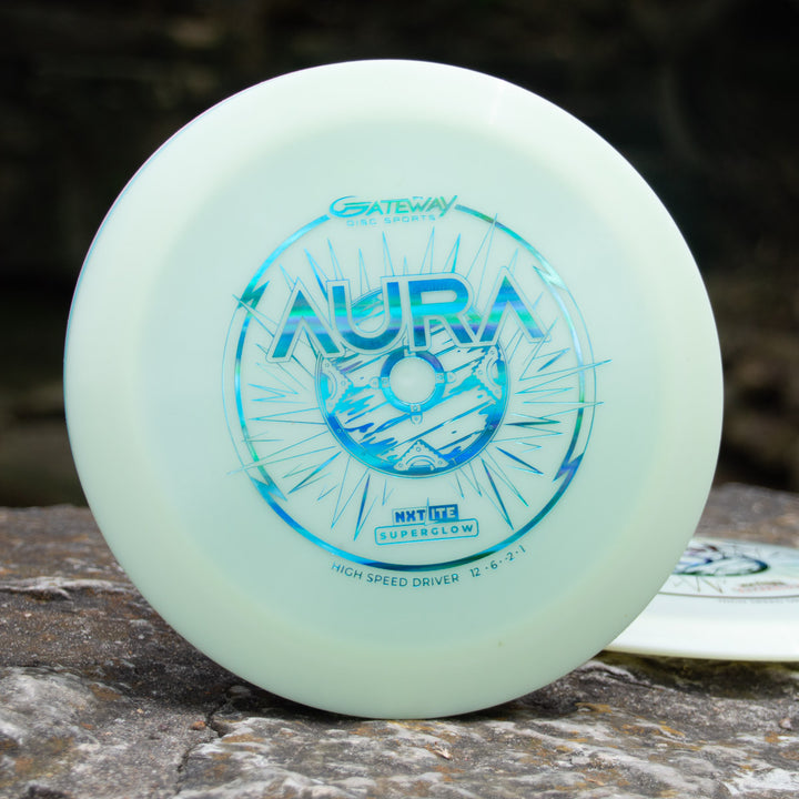 A white "Aura - NXT Lite Superglow" disc golf disc from Gateway Disc Sports with blue graphics rests on a rock—this premium driver is celebrated for its smooth glide.