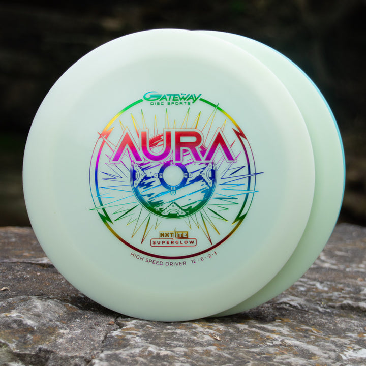 The Aura - NXT Lite Superglow from Gateway Disc Sports is a white top-tier driver with a vibrant logo, shown outdoors on a rock—ideal for smooth hyzer flips and celebrated as a true glide machine.