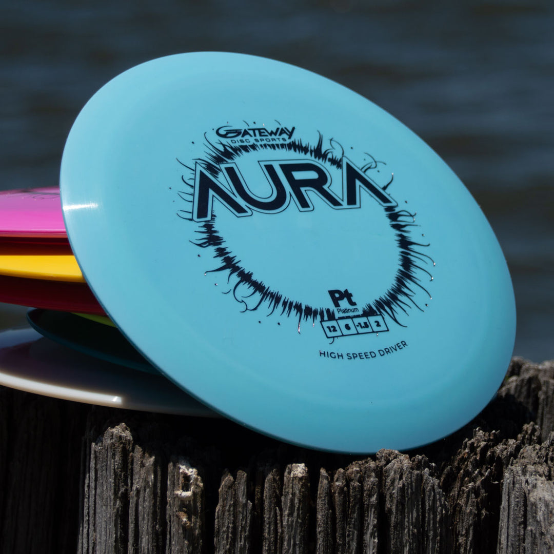 A stack of colorful disc golf discs sits on a weathered wooden post by the water, topped with the blue Aura - Platinum—ideal for long flights and effortless turnover throws.