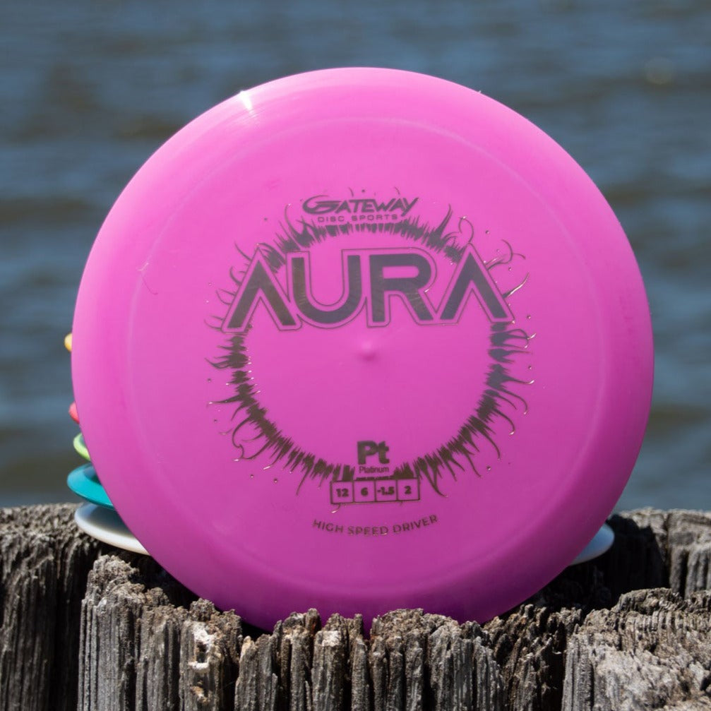 A pink Aura - Platinum disc golf disc by Gateway stands upright on a weathered wooden post near water, ready for easy turnover and long flights.