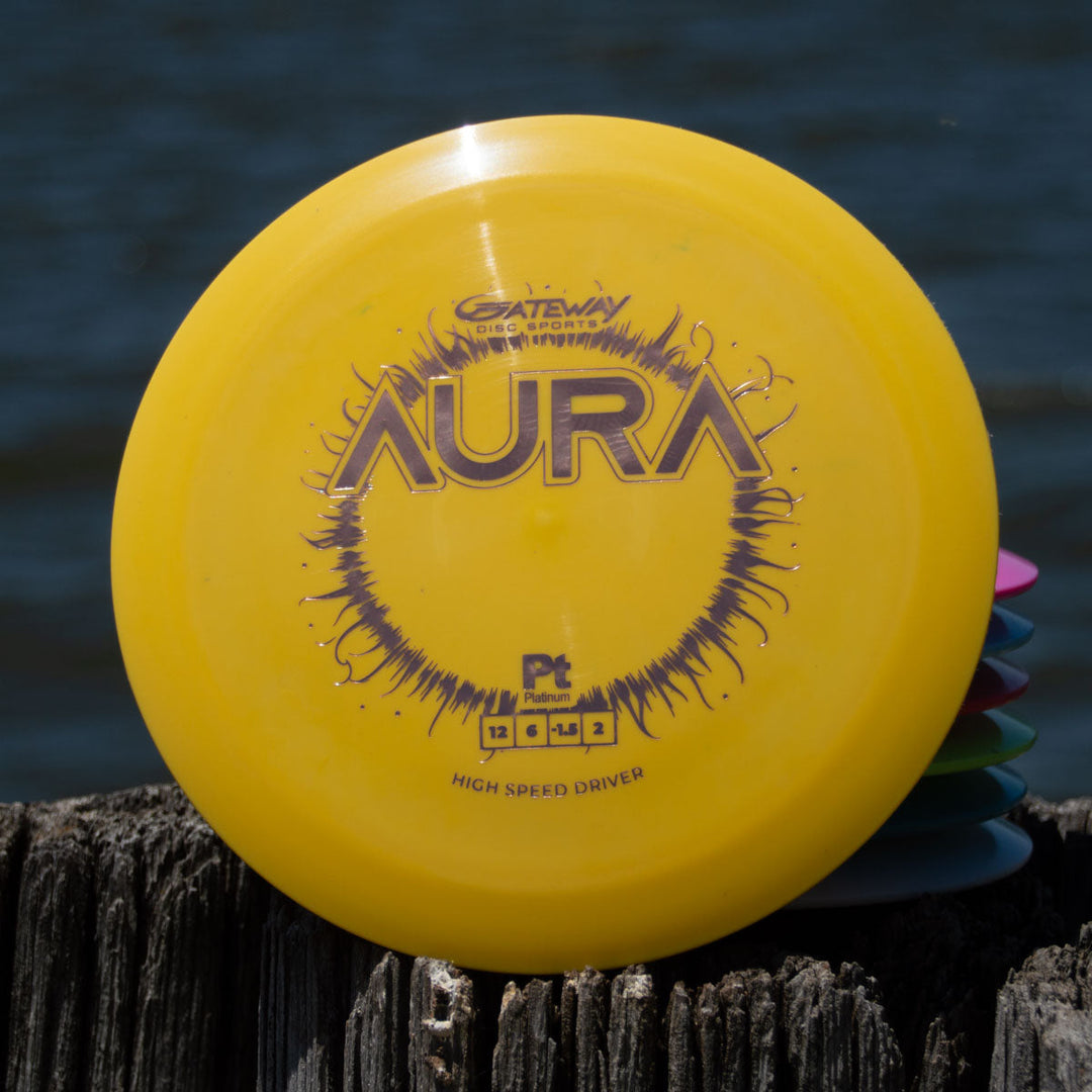 A yellow Aura - Platinum high speed disc golf disc rests on a wooden post by the water, offering easy turnover and long flights, with other discs partially visible in the background.