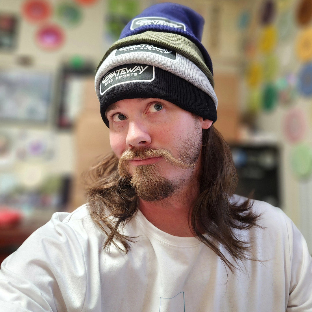 A person with long hair and a curled mustache poses indoors wearing three beanies, including the Gateway Patch Beanie, stacked on their head.