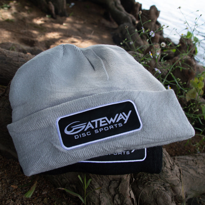 A Gateway Patch Beanie in gray rests atop a black knit cap with a similar Gateway logo, positioned on the ground among tree roots and small plants.