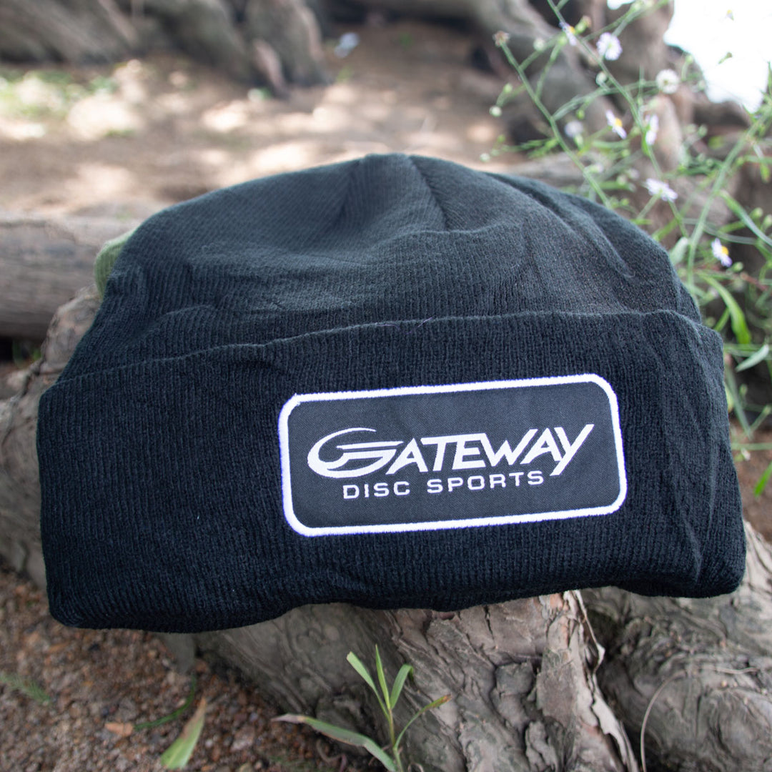 The Gateway Patch Beanie is a black knit sock cap featuring a "Gateway Disc Sports" logo patch, shown outdoors on a tree trunk—ideal for any tournament player's pack.