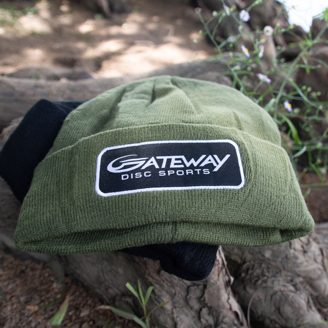 The Gateway Patch Beanie, an olive green knit cap with a black and white "Gateway Disc Sports" patch, sits on a log outdoors—ideal for a tournament players pack with greenery in the background.
