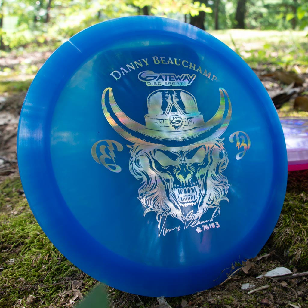 The Danny Beauchamp Diablo - Diamond is a blue overstable fairway driver disc with a skull in a cowboy hat and €œDanny Beauchamp€� on the front, pictured on moss in the woods€”ideal for confident distance flex shots.