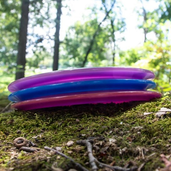 The Danny Beauchamp Diablo - Diamond disc golf driver rests on mossy ground in a sunlit forest, its vibrant colors standing out.