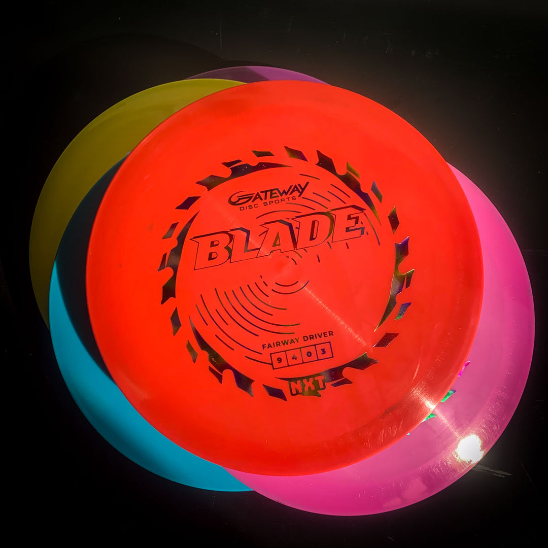 Three colorful disc golf discs—red, yellow, and pink—are stacked. The top red disc is labeled "Blade - NXT," an overstable fairway driver designed for steady resistance during forehand throws.