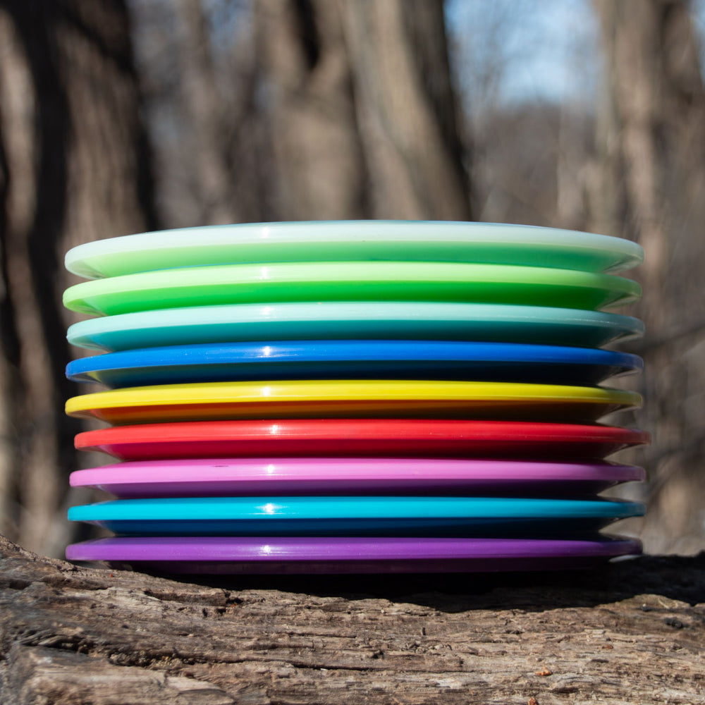 A Blade - NXT overstable fairway driver, stacked with colorful plastic discs, rests on a tree trunk outdoors against a backdrop of blurred trees.