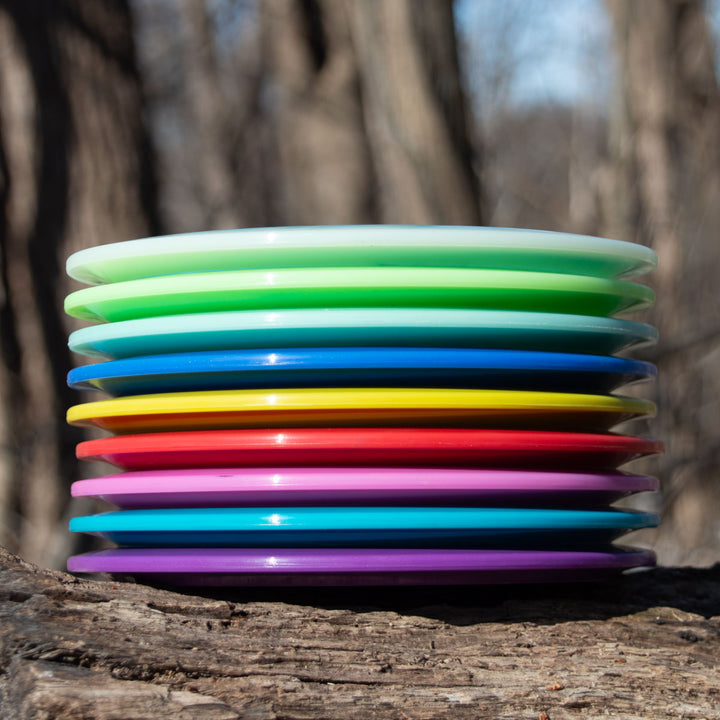 A Blade - NXT overstable fairway driver, stacked with colorful plastic discs, rests on a tree trunk outdoors against a backdrop of blurred trees.
