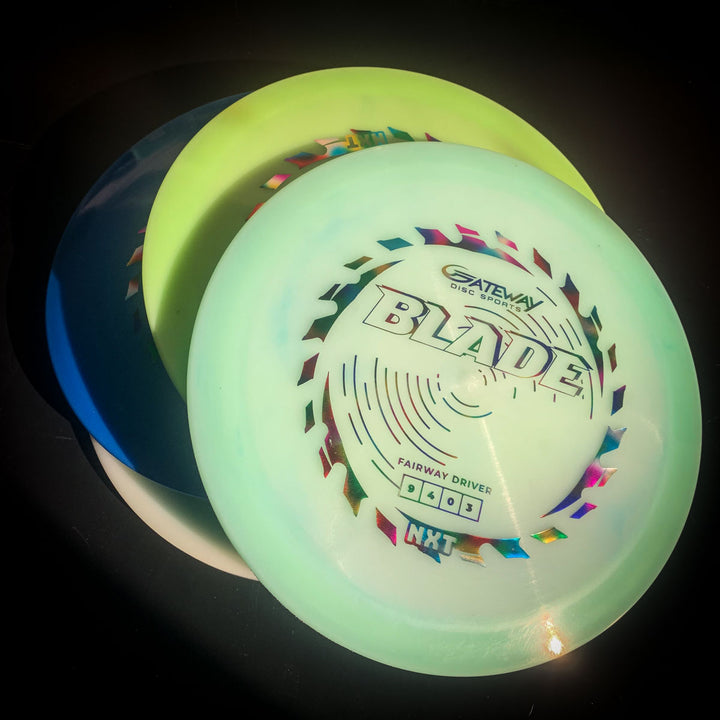 Three overlapping disc golf discs, with the top disc labeled "Blade - NXT" in colorful text, showcased as an overstable fairway driver ideal for steady resistance and controlled forehand shots against a dark background.