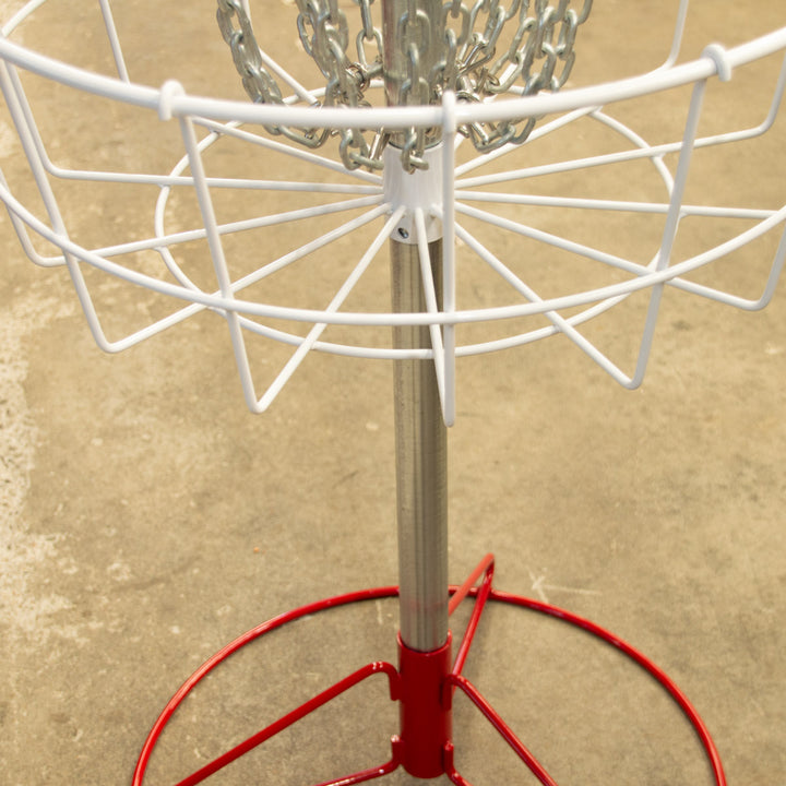 A close-up of the Bullseye™  Portable Disc Golf Basket, showcasing its metal chains and red-and-white frame on concrete—perfect as a disc golf putting trainer for focused practice.