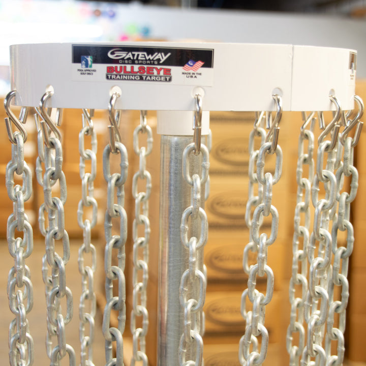 Close-up of a Bullseye™  Portable Disc Golf Basket labeled "Bullseye Training Target," with metal chains hanging from hooks to improve putting. Blurred brown boxes appear in the background.