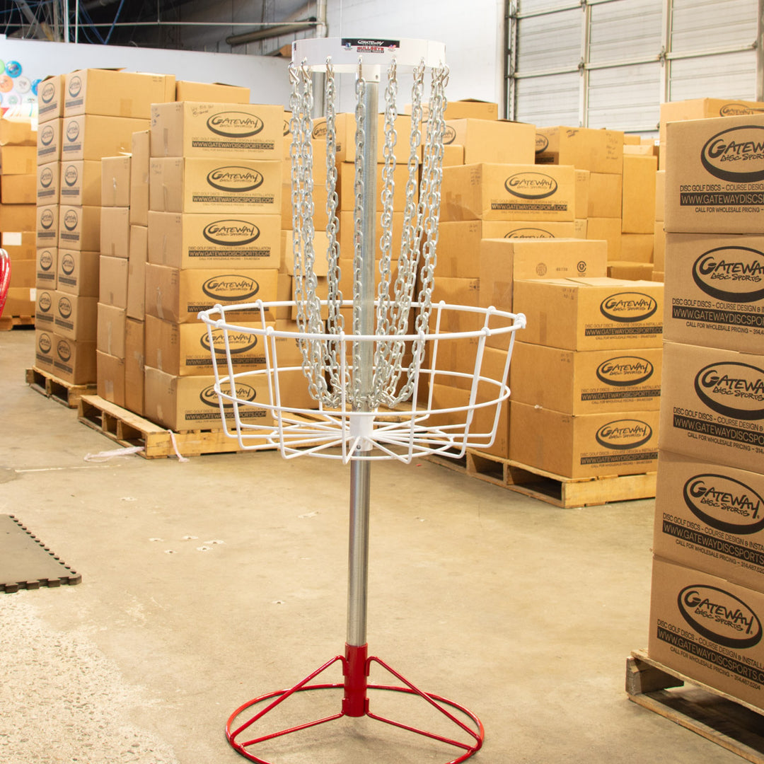 A Bullseye™  Portable Disc Golf Basket, perfect for practice or training, stands in a warehouse surrounded by stacked cardboard boxes on pallets.