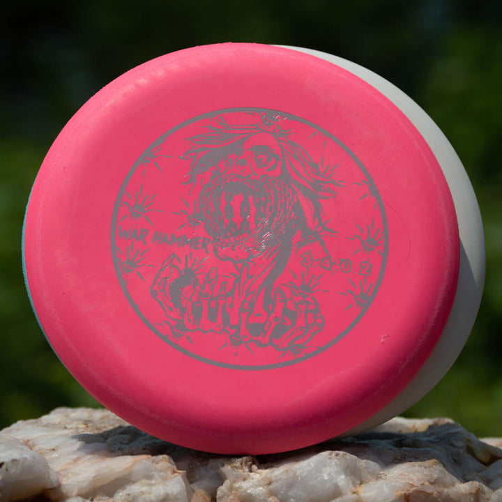 A pink and white Black Zombie Warhammer - ZOMBIE — SSSSS disc, the softest beadless putter, with a metallic roaring monster and claws illustration, rests on a rock amid greenery. A true point-and-shoot disc golf putter.
