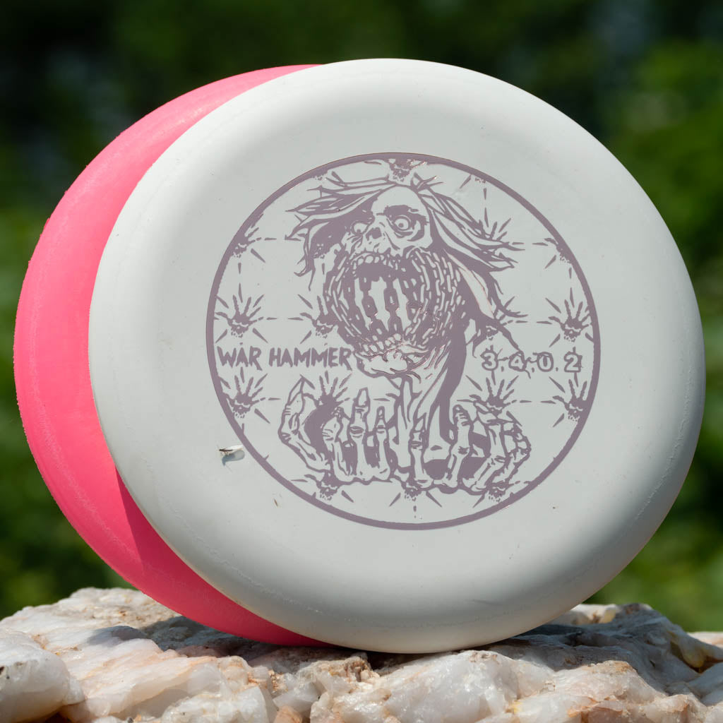 A white beadless Black Zombie Warhammer - ZOMBIE — SSSSS putter disc golf disc featuring a gray skull design and flight numbers 3, 4, 0, 2 leans against a pink disc on a rock outdoors.