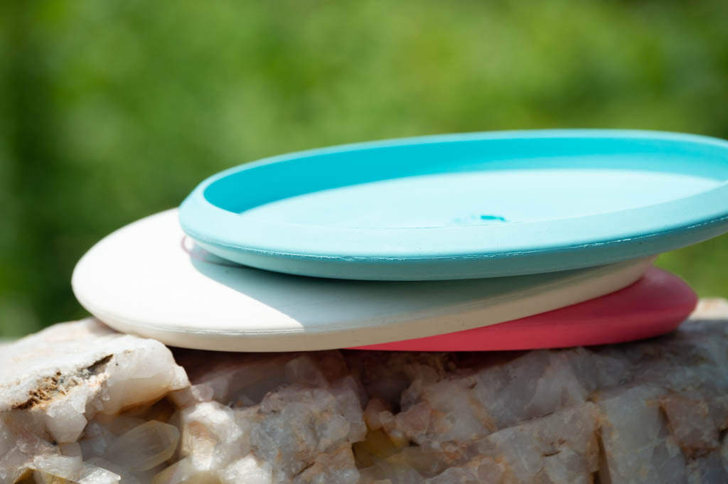 Three plastic Black Zombie Warhammer - ZOMBIE — SSSSS discs—turquoise, white, and pink—are stacked on a rock with a blurred green background, highlighting this soft beadless putter for reliable point-and-shoot accuracy.