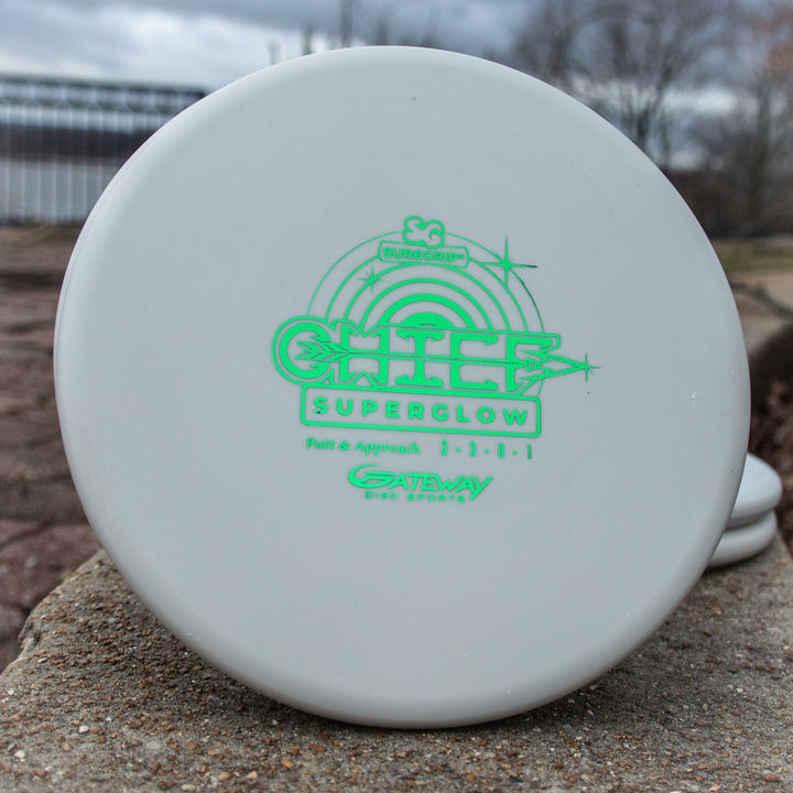 A white Chief - Suregrip Superglow putter with green lettering and a beaded rim stands upright on a stone surface outdoors.
