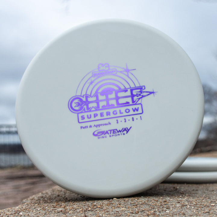 A white low-profile Chief - Suregrip Superglow putter with purple branding sits on a stone surface outdoors.