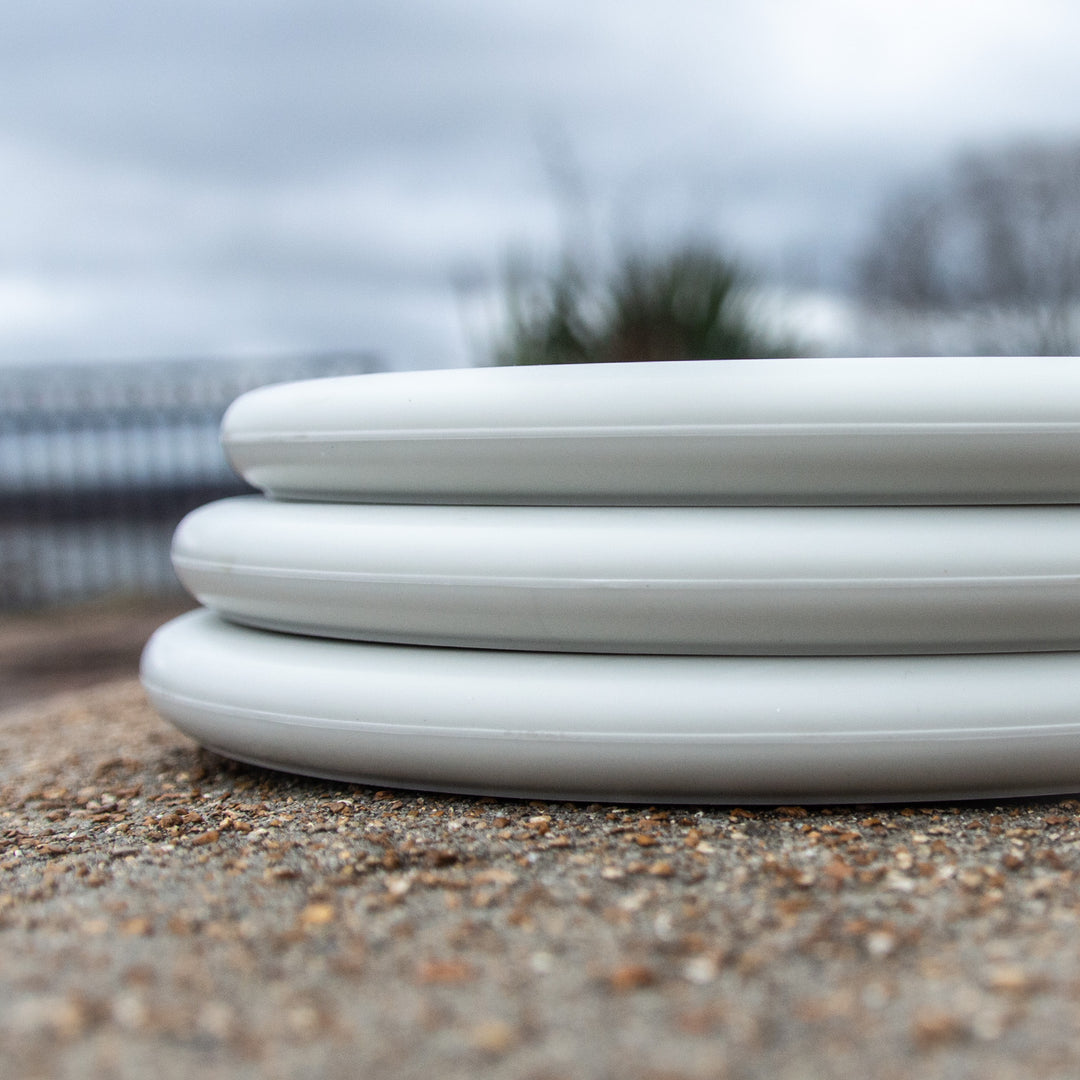 Three stacked Chief Suregrip Superglow plates with a subtle beaded rim rest on a rough, speckled concrete surface outdoors, set against a blurred background of overcast sky and greenery.