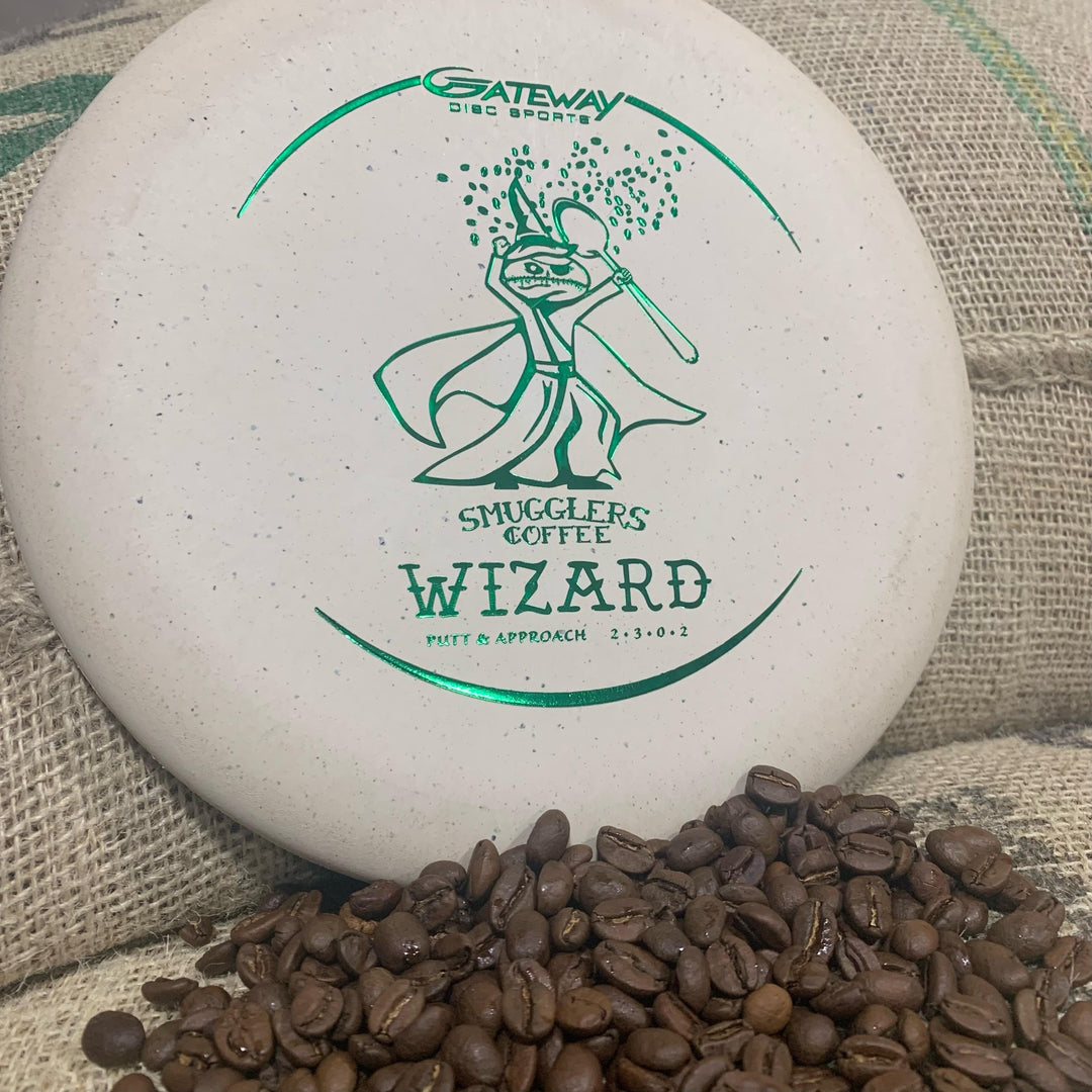 A white Wizard—Spoon Smugglers Coffee Special Blend putter with green graphics and "Smugglers Coffee" text sits by coffee beans and burlap, honoring the bold flavors of Smugglers Coffee's espresso blend.