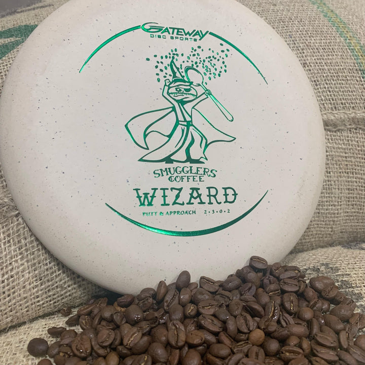 A white Wizard—Spoon Smugglers Coffee Special Blend putter with green graphics and "Smugglers Coffee" text sits by coffee beans and burlap, honoring the bold flavors of Smugglers Coffee's espresso blend.
