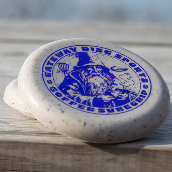 A Coffee Minis disc golf putter with a speckled design, blue wizard graphic, and the text "Gateway Disc Sports," "Warlock," and "GOGFBB Sure Grip," evokes Voodoo putter mystique as it rests on a wooden surface.