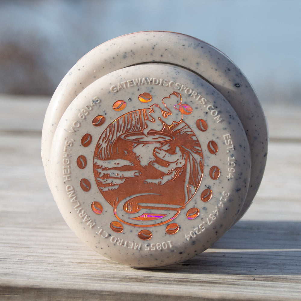 A close-up of a speckled Coffee Minis disc golf putter with an orange stamp, resting on a wooden surface outdoors—ideal for fans of coffee-themed putters.