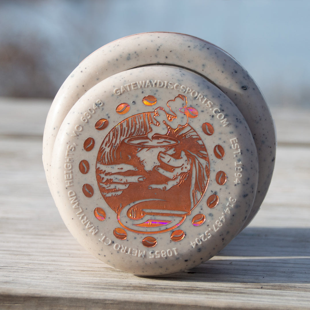 A close-up of a speckled Coffee Minis disc golf putter with an orange stamp, resting on a wooden surface outdoors—ideal for fans of coffee-themed putters.