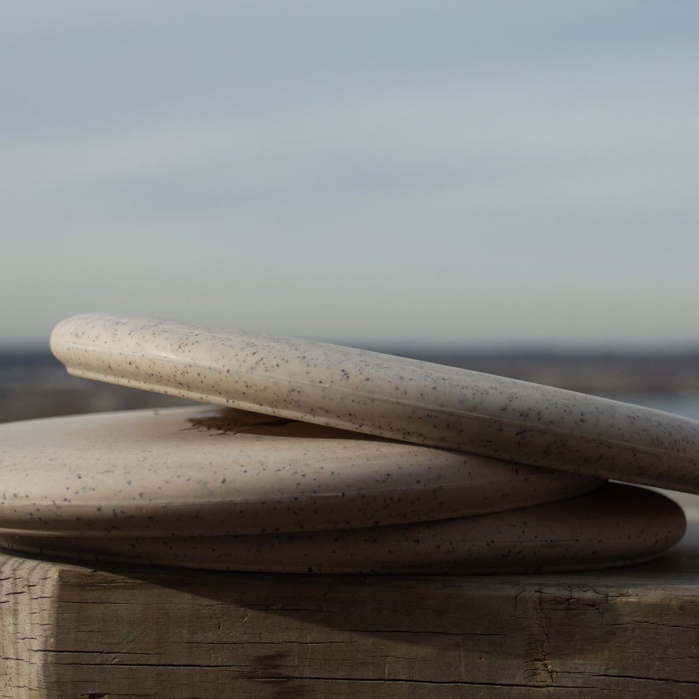 Two speckled ceramic discs, stacked like a stable Wizard Hemp - Air Dude putter, rest on a weathered wooden surface with a blurred outdoor background.