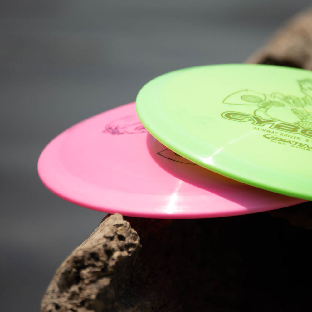 The Cyborg - Diamond flying discs, one pink and one neon green fairway driver, rest on rough textured rock with a blurred background, ready to glide through the air.