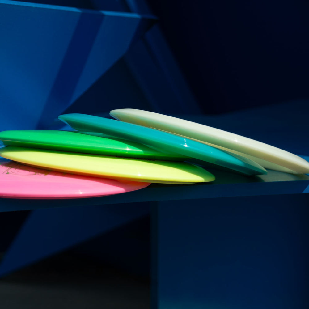 A stack of five Cyborg - Platinum frisbees in pink, yellow, green, blue, and white rests on a blue geometric surface—each a beginner-friendly fairway driver with exceptional glide.