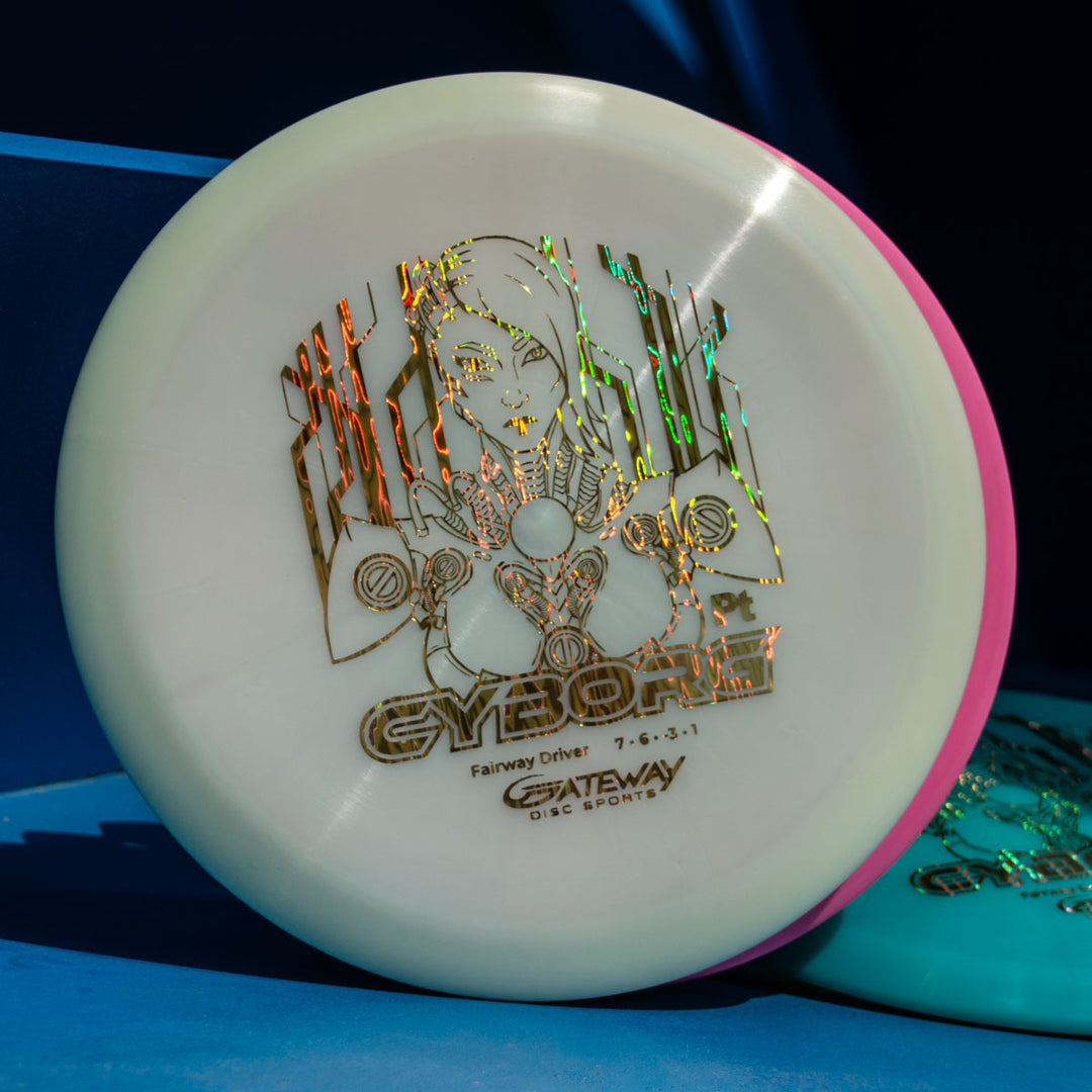 A white Cyborg - Platinum disc golf driver with a holographic cyborg graphic and "CYBORG Fairway Driver 7 6 -3 1 GATEWAY DISC SPORTS" text, known for exceptional glide, rests against a blue background.