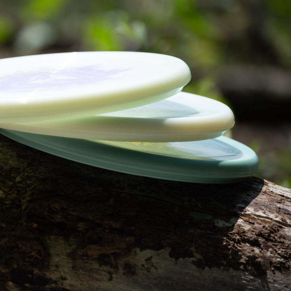 Three plastic discs, including the overstable Devilhawk - Platinum putter with a Thumb Track, are stacked on a tree log against a blurred natural background.