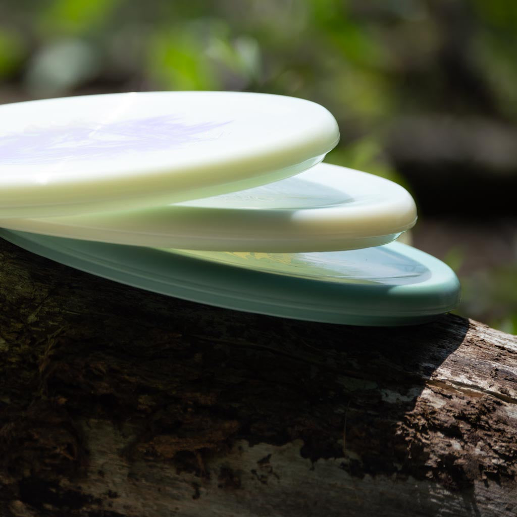 A Devilhawk - NXT Superglow, an overstable putt and approach disc, sits stacked with two other plastic discs on a fallen tree branch outdoors in natural light.