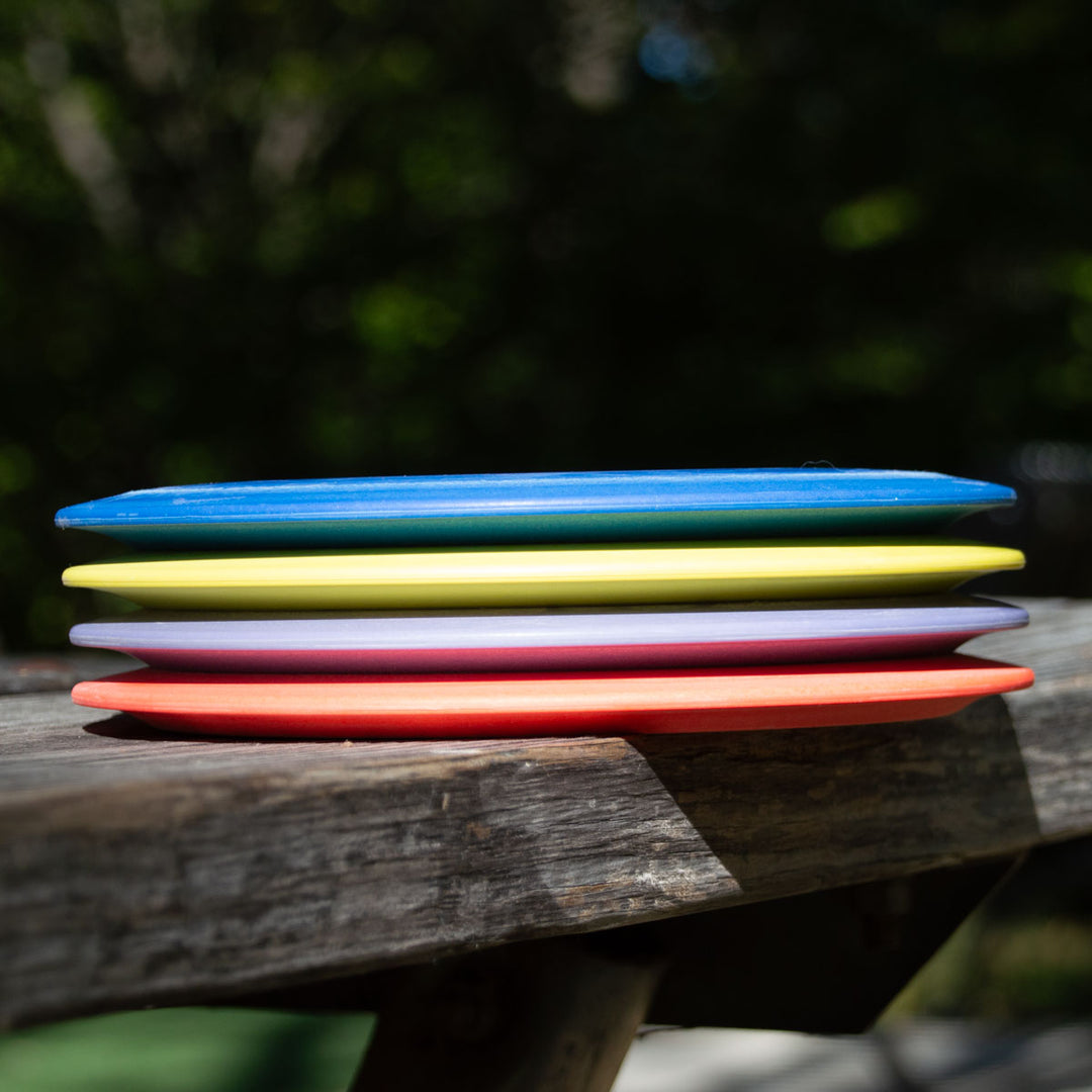 A stack of four Diablo Lightweight - SureGrip discs in blue, green, pink, and red sits on a wooden bench outdoors with a blurred green background—perfect as control drivers or for distance flex shots.