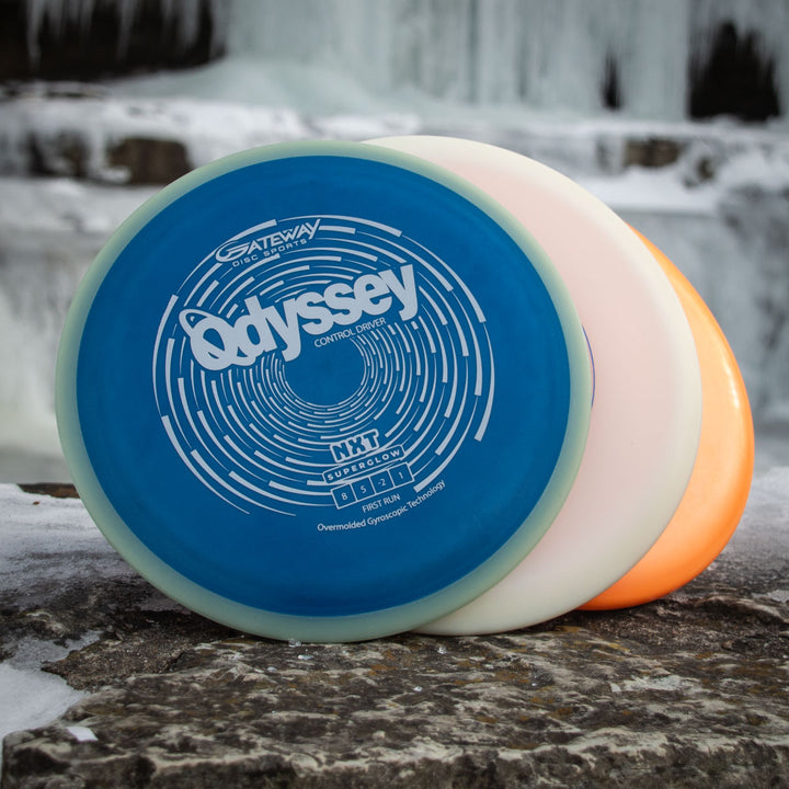 Three disc golf discs, including a blue "Odyssey - NXT Superglow," are propped up on a rock with ice and a waterfall in the background—an ideal setting to imagine the Odyssey Story by David McCormack unfolding.