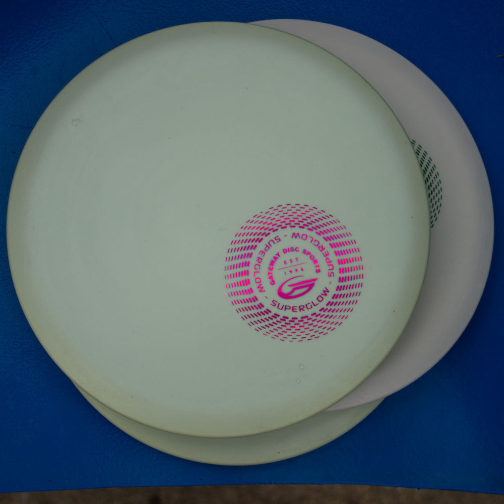 Two stacked Element - Suregrip Superglow discs, white and off-white, with a pink logo on top, rest on a blue surface—ideal for players seeking great glide and a straight flight path.