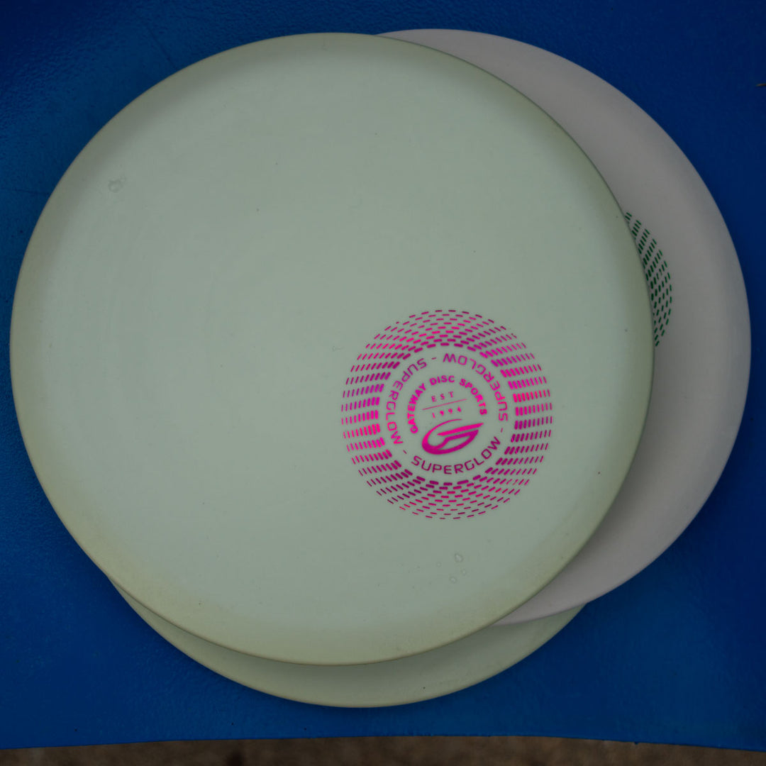 Two stacked Element - Suregrip Superglow discs, white and off-white, with a pink logo on top, rest on a blue surface—ideal for players seeking great glide and a straight flight path.