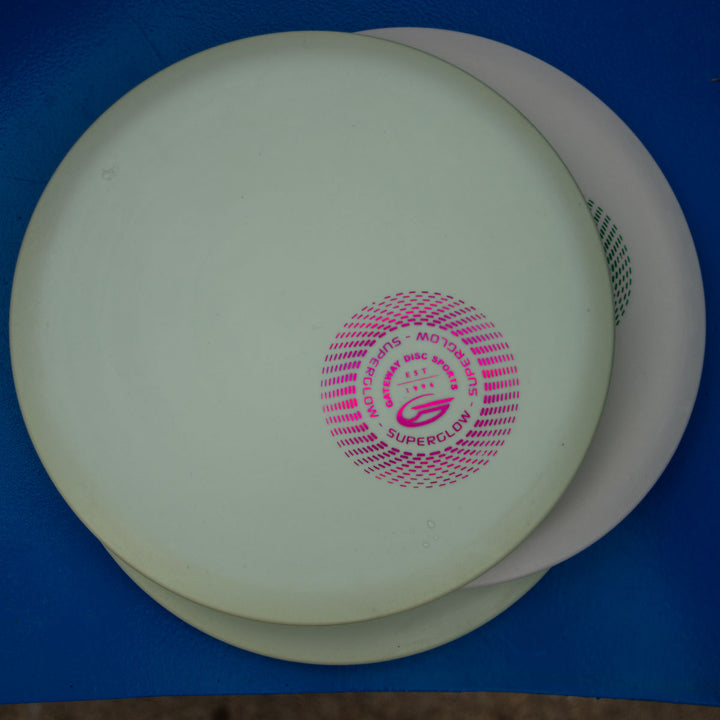 Two stacked Element - Suregrip Superglow discs, white and off-white, with a pink logo on top, rest on a blue surface—ideal for players seeking great glide and a straight flight path.