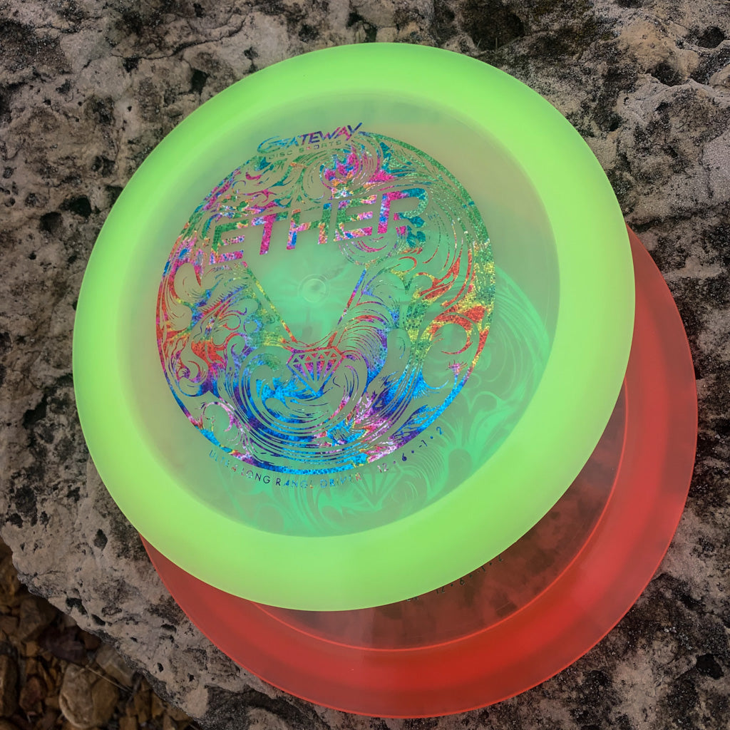 Two vibrant Ether - Diamond disc golf discs—one green, one red—featuring intricate designs rest on a textured rock. Both are PDGA approved distance drivers, expertly crafted for optimal glide and standout performance.