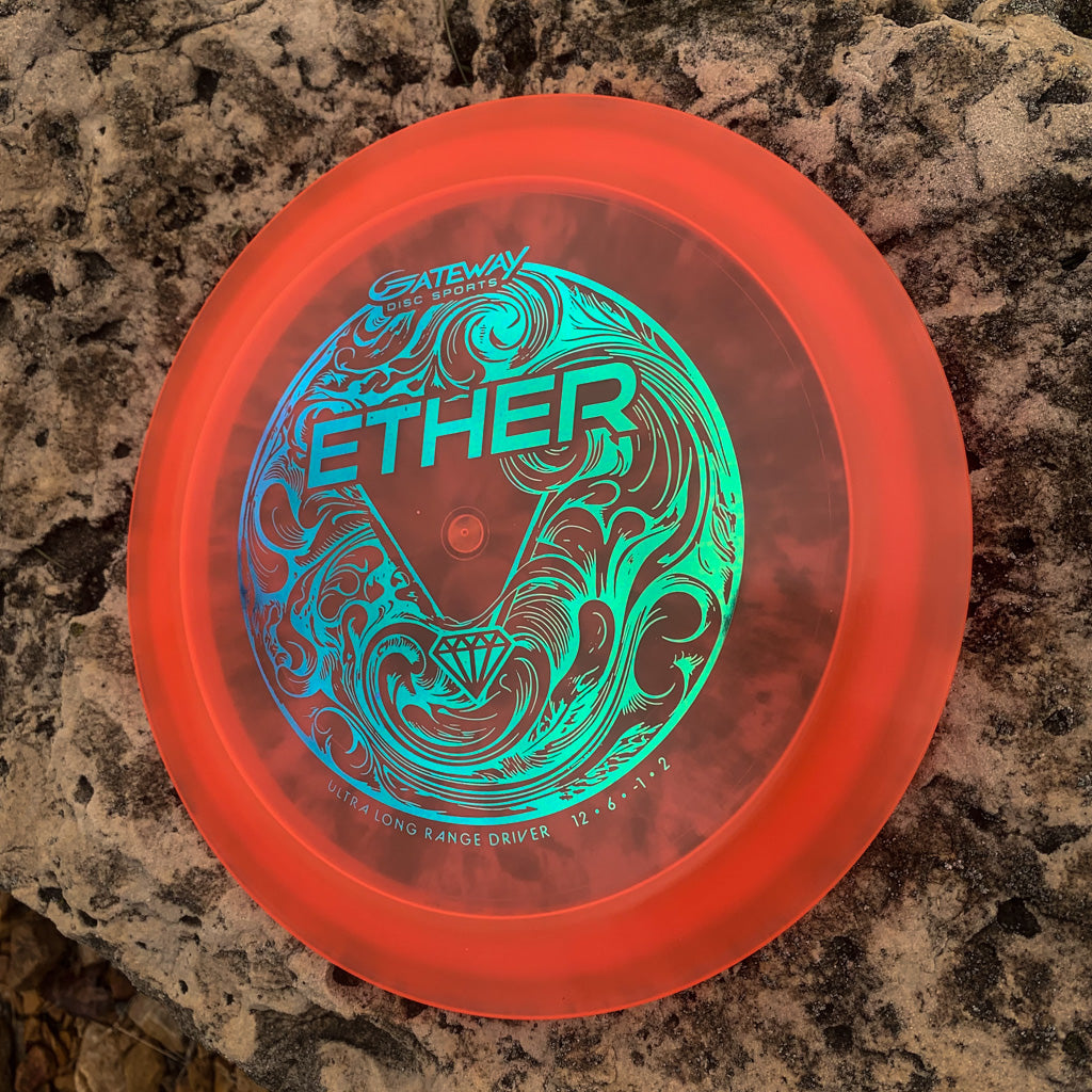 A red Ether - Diamond distance driver by Gateway, featuring aqua blue artwork and PDGA approval, lies on a textured rock surface.