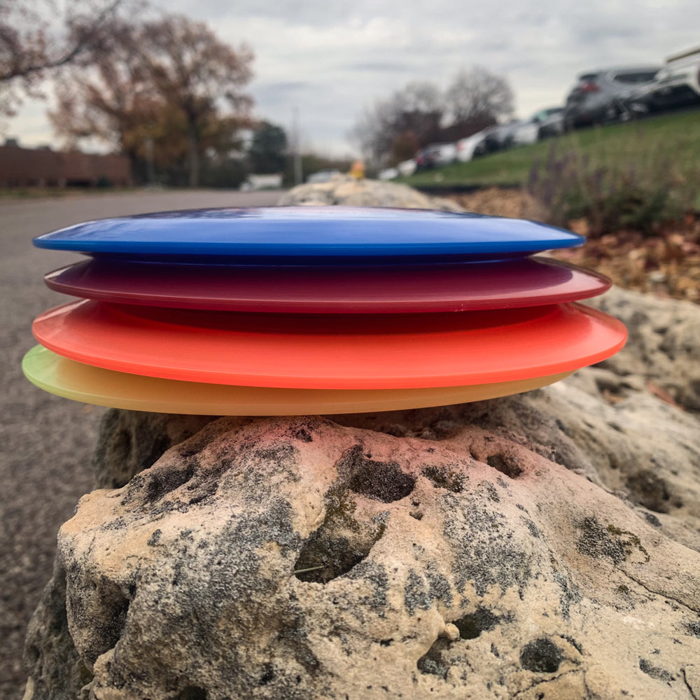 The Ether - Diamond, a vibrant PDGA approved disc golf disc, is stacked on a large rock with a road, cars, and trees in the cloudy background.