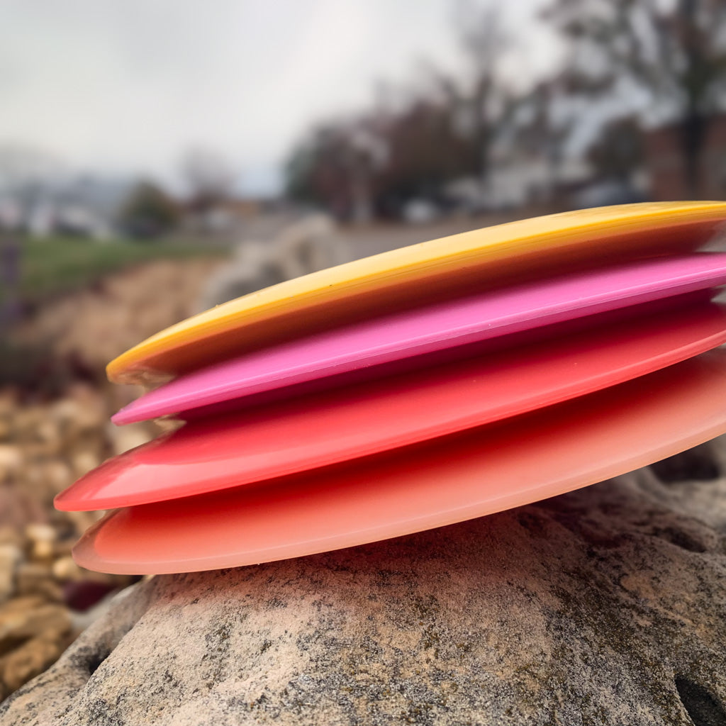 Four colorful Ether - NXT (Nylon Xtra Tuff) disc golf discs—yellow, pink, red, and orange—stacked on a rock with a blurred outdoor background. PDGA approved distance drivers made from durable Nylon Xtra Tuff material.