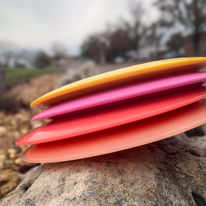 Four colorful Ether - NXT (Nylon Xtra Tuff) disc golf discs—yellow, pink, red, and orange—stacked on a rock with a blurred outdoor background. PDGA approved distance drivers made from durable Nylon Xtra Tuff material.