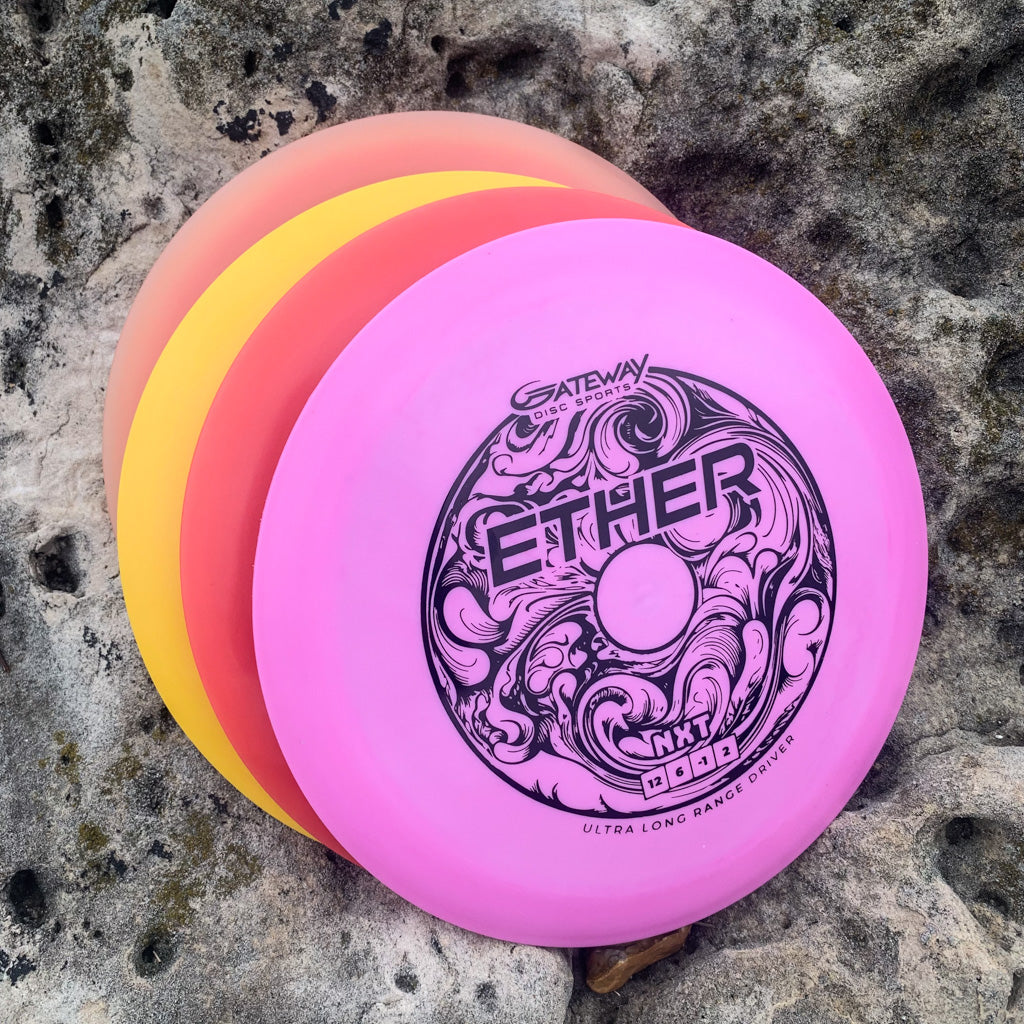 Three disc golf discs—pink, orange, and yellow—are stacked on rocks. The top disc is the Ether - NXT (Nylon Xtra Tuff) distance driver with an intricate design and PDGA approval, known for its exceptional durability.