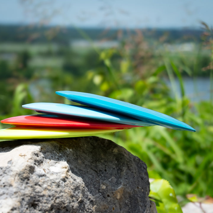 Four Ether - NXT LITE disc golf discs stacked on a rock outdoors, surrounded by greenery and a blurred landscape—ideal for highlighting their unique flight numbers.