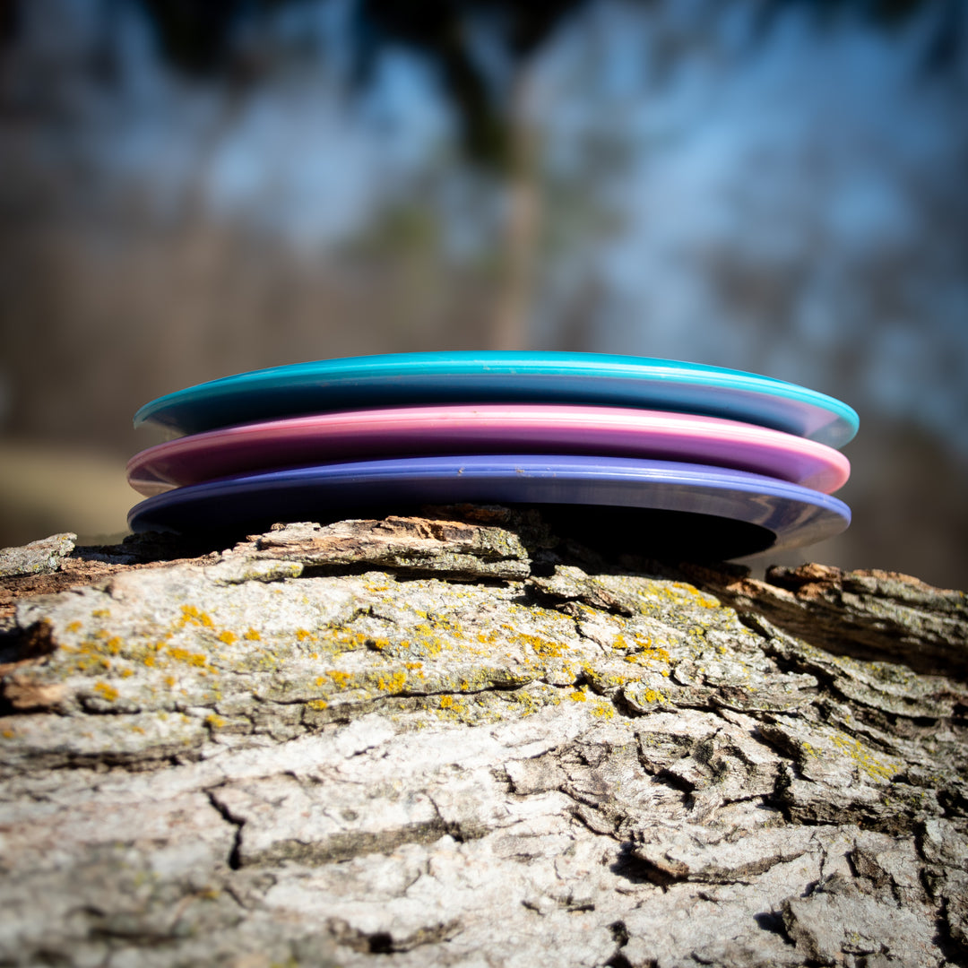 Three Ether - Platinum - New Run disc golf discs, featuring a distance driver with unmatched grip, stacked on a tree trunk outdoors against a blurred natural background.